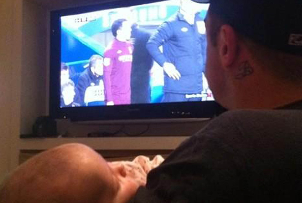 Robbie Williams är nybliven pappa. Här kollar han fotboll med bebisen framför teven. 