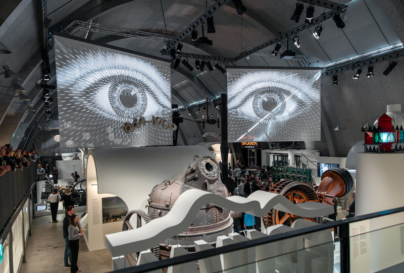 På två skärmar i taket visas med jämna mellanrum det nya audiovisuella verket ”Teknika & neutika” av Try New Things med fokus på människorna och på hur deras liv påverkas av den snabba tekniska utvecklingen. Pressbild. 