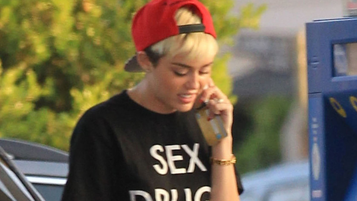 Miley Cyrus möter sommaren i shorts, röd keps och en t-shirt med budskapet: Sex, drugs & Rap.