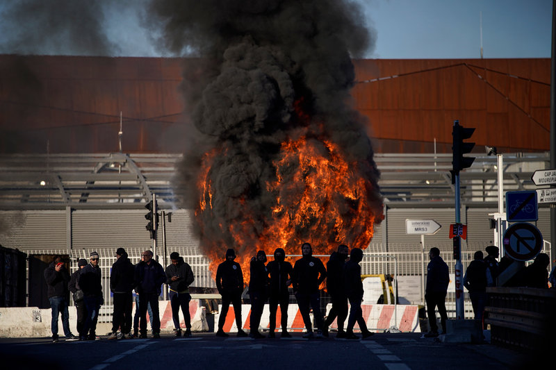 Strejkande hamnarbetare i Marseille har tänt eld på en barrikad som blockerar vägen till hamnen, på onsdagsmorgonen.