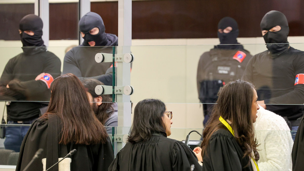 Försvarsadvokaterna talar med de åtalade inför rättegångsstarten i Bryssel, med maskerad polis i bakgrunden.